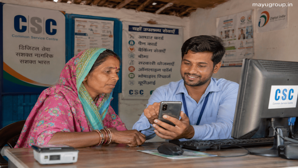 Rural citizen using a smartphone at a Common Service Centre while an operator assists with digital government services in a village office.