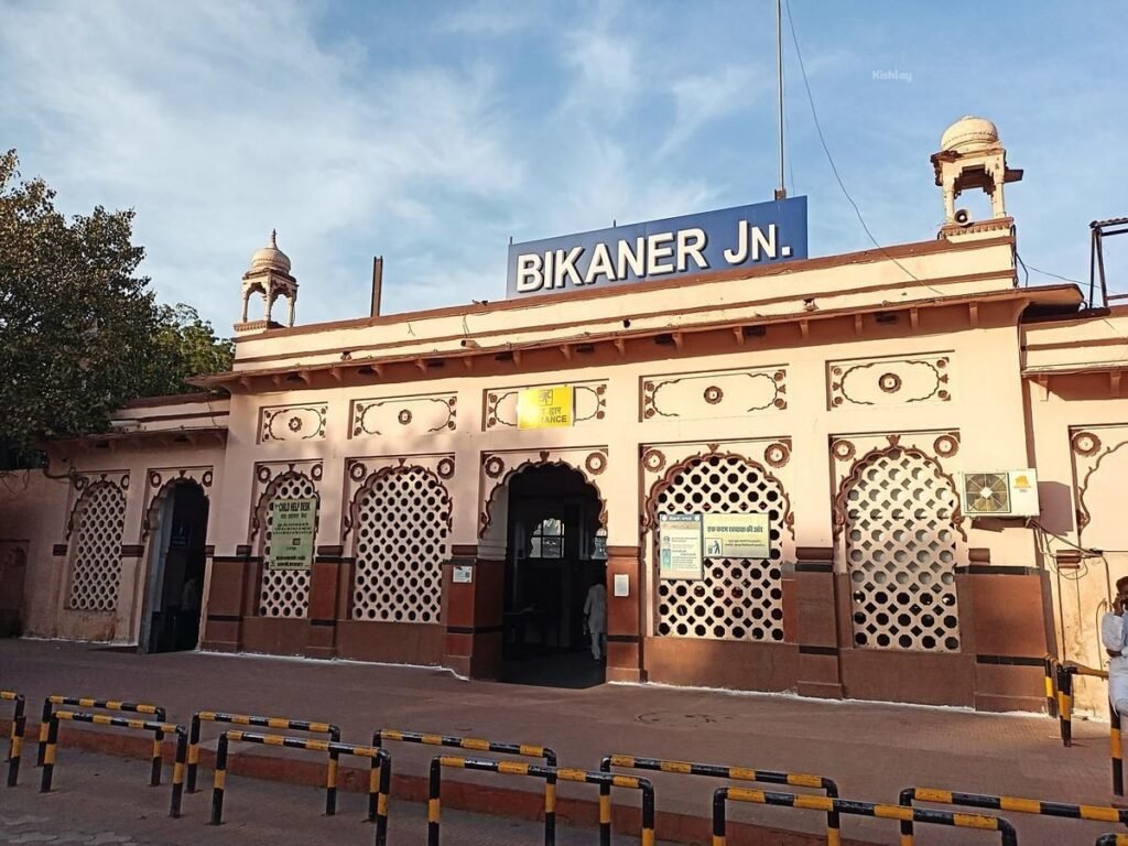 Exterior entrance view of the heritage building at Bikaner Junction Railway Station showing decorative jali lattice screens, arched entryways, small corner chhatris, and a blue signboard displaying Bikaner Jn.