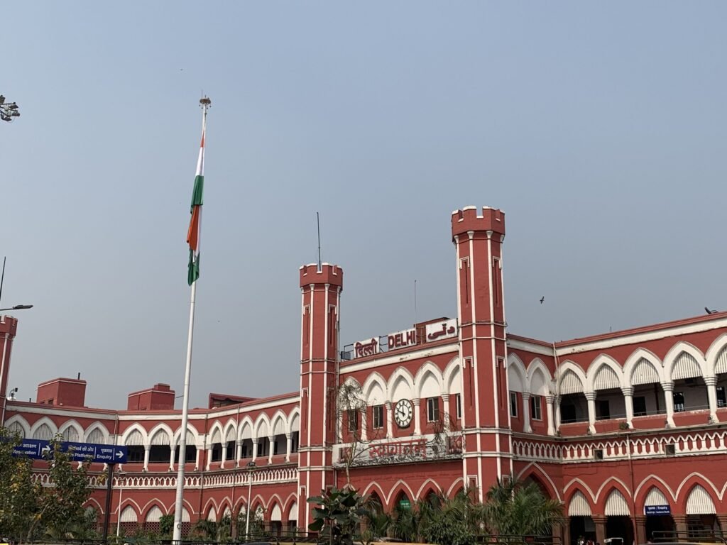 Exterior view of the historic red colonial building of Delhi Junction Railway Station featuring octagonal turrets, Gothic arched arcades, a central clock, and a tall Indian national flag flying in the foreground.