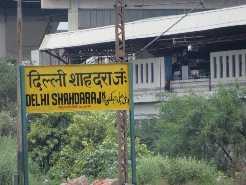 Yellow multilingual station signboard reading Delhi Shahdara Junction in Hindi, English, and Urdu, with an elevated Delhi Metro rail structure and green foliage in the background.