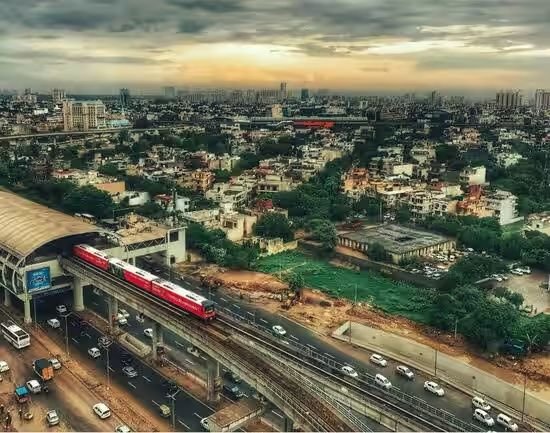 High-angle aerial view of an elevated rapid transit metro line and station in Gurgaon, featuring a red metro train departing the platform above a busy multilane highway with dense urban cityscape in the background.