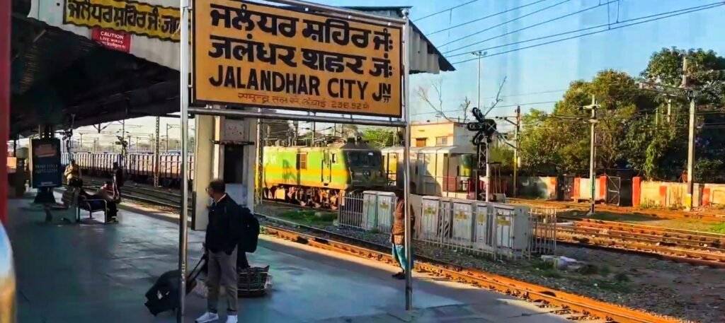 Platform view of Jalandhar City Junction Railway Station showing a yellow multilingual station signboard in Punjabi, Hindi, and English, with passengers waiting and an electric locomotive visible in the background tracks.