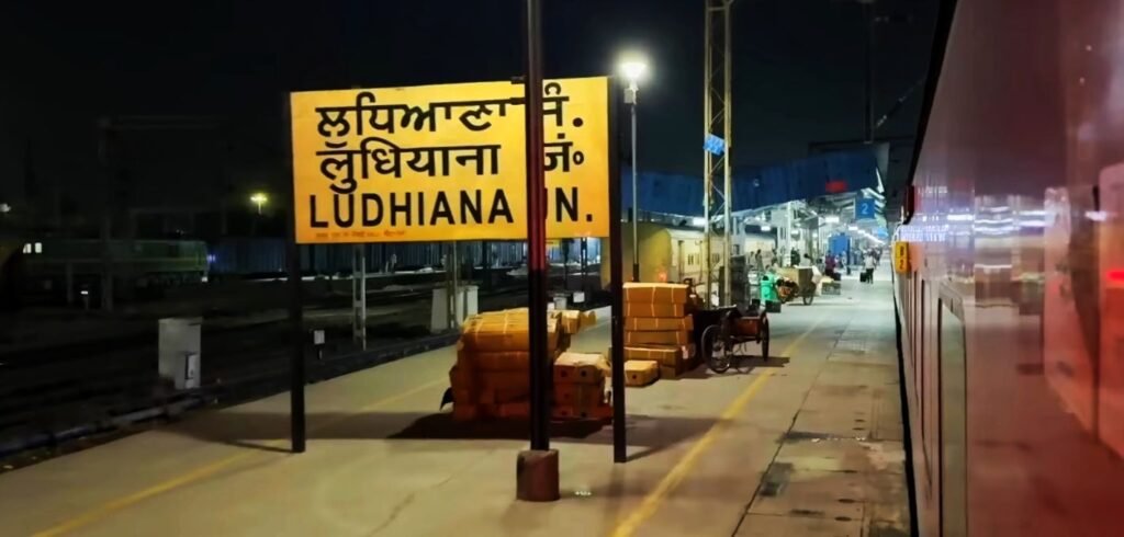 Nighttime platform view at Ludhiana Junction Railway Station featuring an illuminated yellow station signboard in Punjabi, Hindi, and English, with cargo boxes stacked nearby and a train coach on the right.