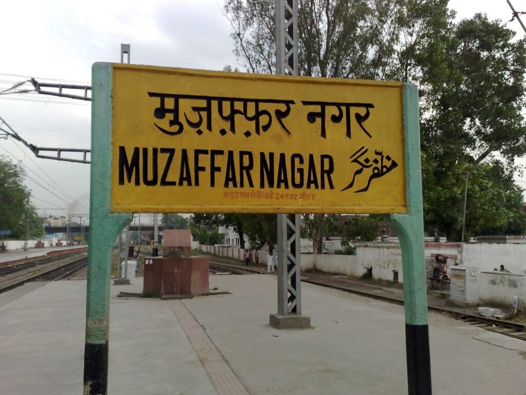 Close-up view of a free-standing yellow station signboard at Muzaffarnagar Railway Station displaying the station name in Hindi, English, and Urdu, supported by teal posts on an open platform with tracks behind it.