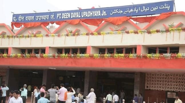Exterior entrance view of Pt. Deen Dayal Upadhyaya Junction railway station building featuring a large blue multilingual sign header, orange fabric drapes, floral garlands, and people gathered out front.