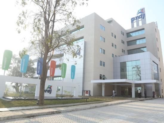 Exterior view of the multi-story Praj Industries Limited corporate office building featuring modern architectural glass facade, rooftop logo signage, paved entrance driveway, and colorful outdoor promotional feather flags set against a bright sky.