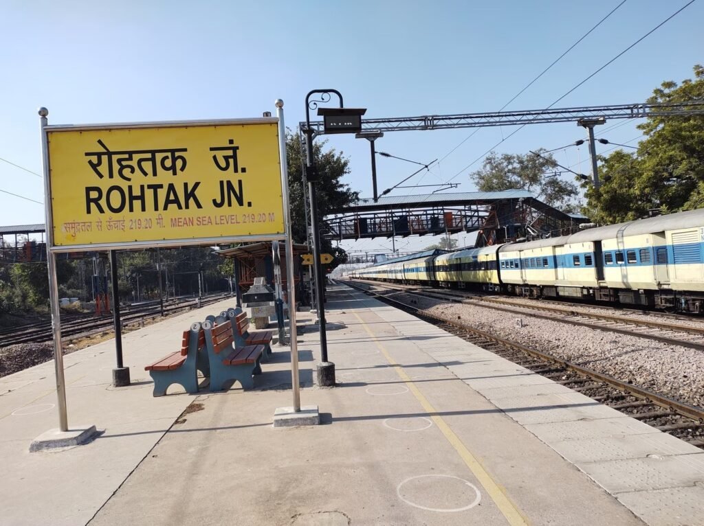 Platform view at Rohtak Junction Railway Station featuring a yellow station signboard in Hindi and English with elevation details, platform seating, overhead electrification infrastructure, and a long passenger train stationary on adjacent tracks.